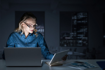 Attractive blonde working on laptop in dark office. Mixed media