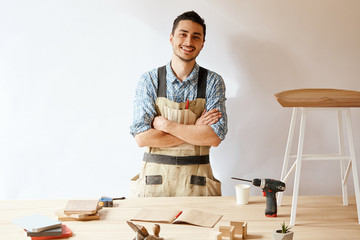 Skilled craftsman in apron looking up from his work and smiling at camera while standing in his...