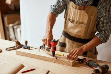 Carpenter doing his job in carpentry workshop, drilling a hole with an electrical drill in workshop. Profession, carpentry, woodwork and people concept.
