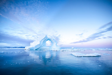 Photogenic and intricate iceberg with a hole under an interesting and colorful sky during sunrise with full moon. Disko bay, Greenland. - obrazy, fototapety, plakaty