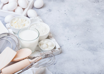 Fototapeta na wymiar Fresh dairy products on white table background. Glass of milk, bowl of flour and cottage cheese and eggs. Box of baking utensils. whisk and spatula in vintage wooden box.Top view. Space for text
