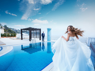 Bride and groom near the swimming pool. Honeymoon wedding the young couple. The woman in a beautiful dress runs to the man in the suit. Luxury resort.