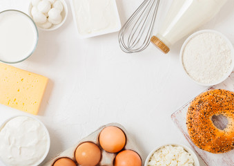 Fresh dairy products on white table background. Glass of milk, bowl of sour cream and cottage cheese and eggs. Fresh baked bagel. Steel whisk. Top view.Space for text