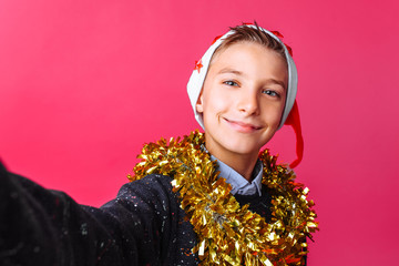 A teenager guy takes a selfie in a Santa hat and tinsel on the neck, on a red background.