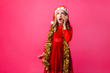 Obraz na płótnie Canvas Portrait of schoolgirl girl in Santa hat and tinsel on neck, emotionally talking on phone with surprised facial expression isolated on red background