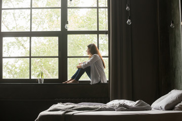 Lonely sad brunette female in casual clothes sitting on window sill barefoot and looking at window...