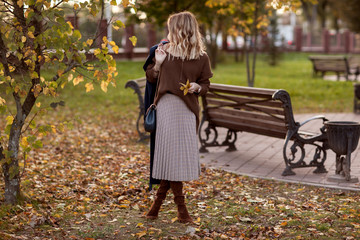 A beautiful fashionable woman is standing in an autumn park in a brown sweater and a long plaid skirt with a bag in her hands.