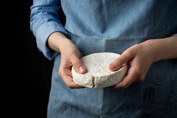 Woman in blue apron holding soft french camembert cheese on dark background