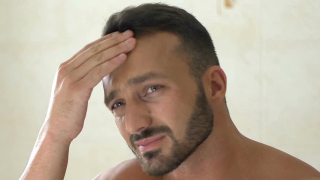 Unhappy man checking wrinkles on face in front of mirror in bathroom, aging