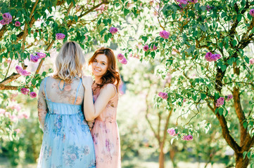 Adorable happy cheerful fabulous twin sisters in different beautiful summer dresses posing outdoor.  Similar cute female models in lilac colorful bushes with bloming flowers in park portrait.  Family.