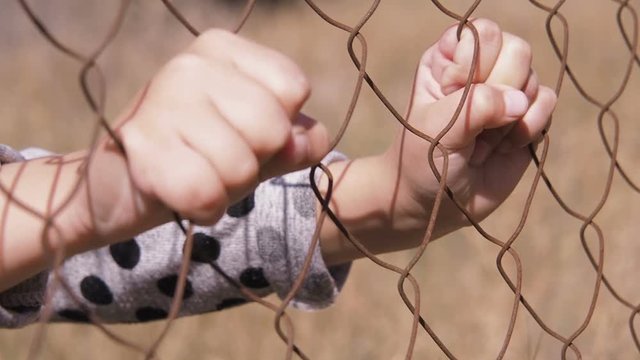 Homeless child. The hands of the child and the iron grid. Refugee camp.