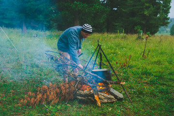 Tourist kindles fire. Traveler is lighting fire in camp. Man puts dry branch in campfire. Active outdoor recreation. Into the wild. Camping in dusk. Atmospheric warm in twilight. Active rest.