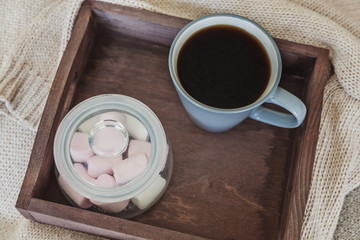 Cup of coffee on rustic wooden tray, sweet marshmallow and warm woolen sweater. Cozy autumn or winter weekend or holidays at home. Fall home decoration with hot drink mug. Hygge morning style concept