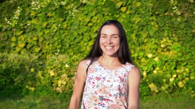 European young woman with long black hair runs away from the camera in slow motion, calling for herself. Attractive girl with dimpled cheeks in a white floral print beckons to follow her. Smiling