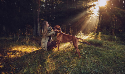 Young smiling woman with dreadlocks in autumn fall forest in the morning sunshine playing with a dog ridgeback