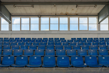 Front view row of blue plastic seats on rough concrete step floor in old vintage dirty concrete structural stadium with windows frame background without people.  