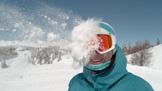 SLOW MOTION, CLOSE UP: Excited girl wearing ski goggles gets hit in the head by snowball. Unrecognizable person trows a ball of fresh powder snow at a smiling young woman on active winter holiday.