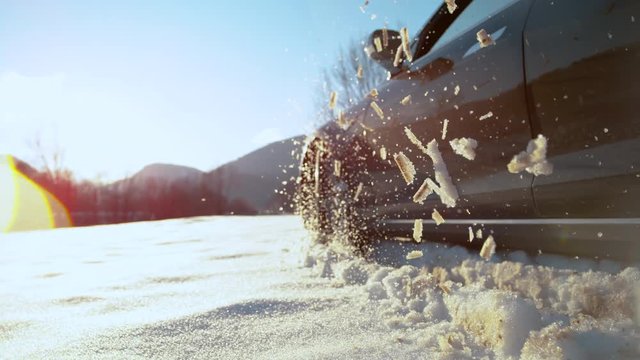 SLOW MOTION, CLOSE UP, LENS FLARE: Bright winter snow shining on black car driving in the fresh snow. Cinematic shot of wet snow flying off powerful car's tyres. Tyre losing traction in slippery snow.
