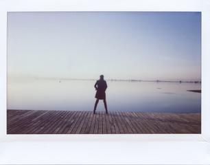 Polaroid back view of woman standing on pier in morning.