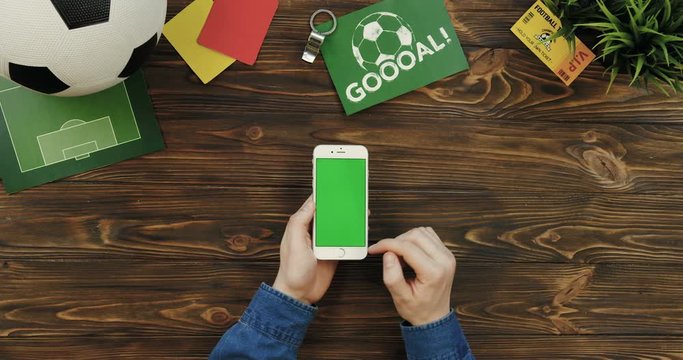 Top view on the white smartphone with chroma key screen being vertically on the wooden table and male hands scrolling and taping on it. Football ball, goal card and fan ticket lying beside. Green