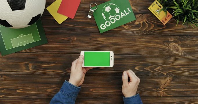 Top view on the white smartphone with chroma key screen being horizontally on the wooden table and male hands scrolling and taping on it. Football ball, goal card and fan ticket lying beside. Green