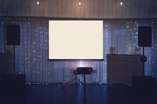 Illuminated banquette hall stage with a big white screen and acoustic systems. Equipment for video and audio projection at a festive event. Banquette stage with a board for projection in the centre