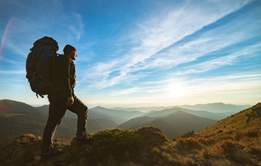 The man standing with a camping backpack on the rock with a picturesque sunset
