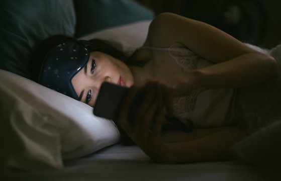 Young woman using phone resting on bed at night.