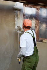 Worker in overalls and cap painting primer on wall with roller before plastering. Facade thermal...