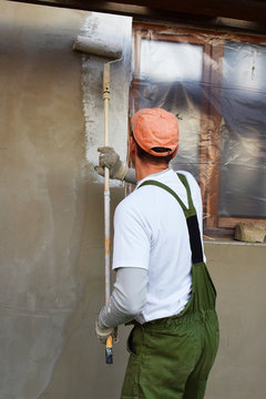 Worker in overalls and cap painting primer on wall with roller before plastering. Facade thermal insulation and painting works during exterior renovation. Home improvement and DIY concept.