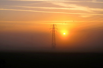 electricity pylon with the sun rising next to it there is mist on the field in the fore ground and the sky has a deep orange glow