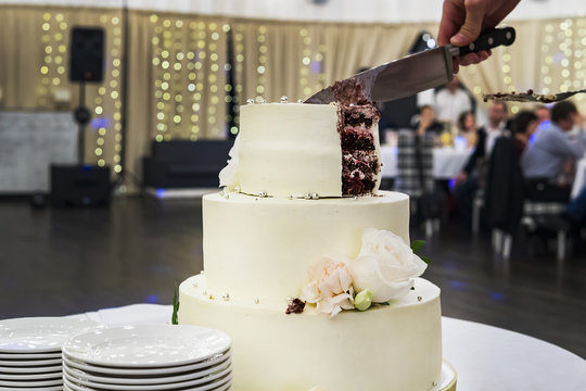 Cutting the first tier of white mastic wedding cake in banquet hall background. Front view at table with a beautiful wedding cake and side plates for serving guests. Dividing wedding cake into parts.