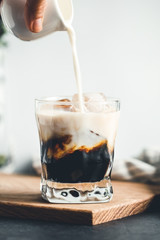 Pouring milk in a glass with espresso and iced cubes in a modern kitchen in the morning.