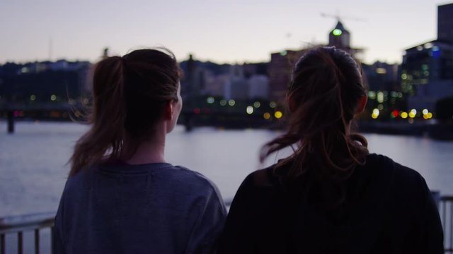 Tracking Shot Of Friends Walking Down Spiral Ramp, At Sun Down, City Lights Across River In Background, Slow Motion