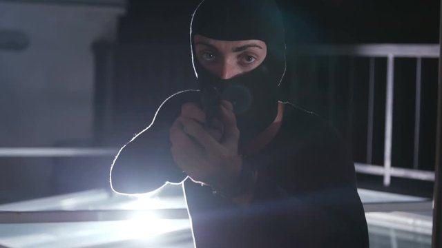 Caucasian criminal in black balaclava turning gun and looking directly at camera. Masked bandit going to commit crime. Lifestyle. Night time.