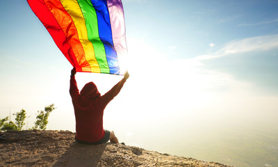 man sitting on mountain top waving rainbow LGBT symbol flag in bright sunlight blue sky