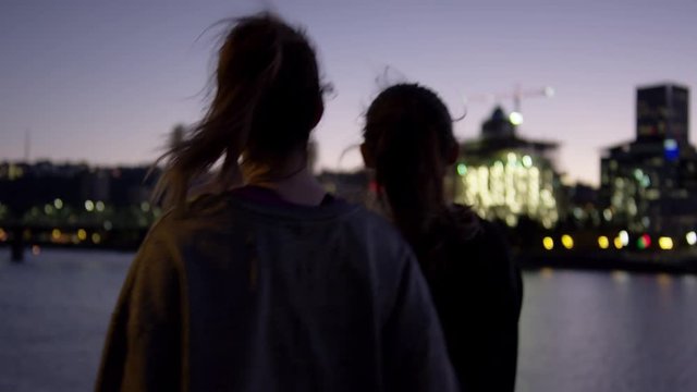 Friends Walk Along River At Night, Woman Holds Up A Peace Sign Briefly, City In Background