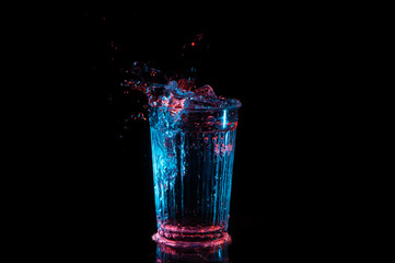 Liquid splashing out of a vintage lined glass under blue and red lights isolated on a black...