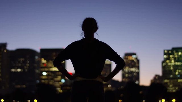Young Woman Put Her Hands On Her Hips In Power Pose, Looks Out At City Across River, Camera Tilts From Feet Up