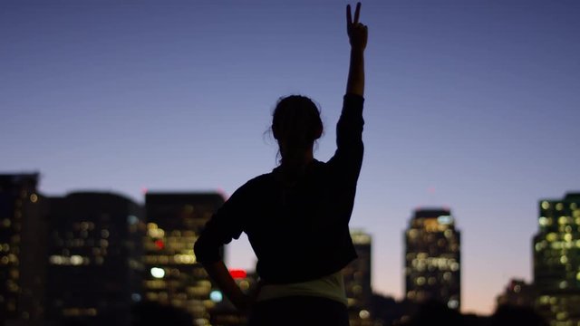 Young Woman Puts Her Hands On Hips, Then Raise Hers Hands In Air, Makes Peace Signs, City In Background