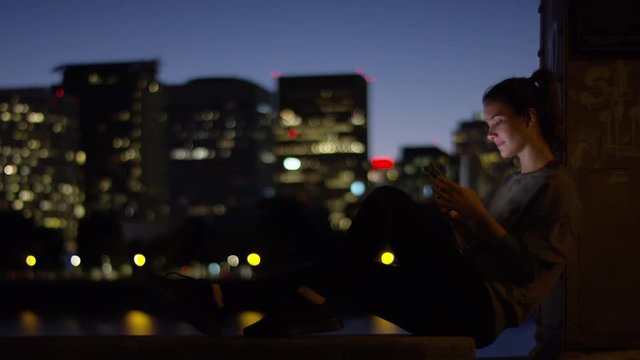 Happy Young Woman Rests After Workout, Plays On Smartphone By River, City Lights In Background