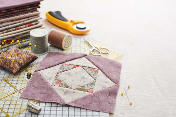 Patchwork block on craft mat, stack of fabric, sewing accessories on white wooden surface