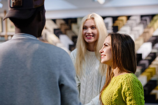 Three multi ethnic people students talking, looks at each other indoor at frames store, smiling. Young friendly black skinned guy and two european girls choosing a gift.