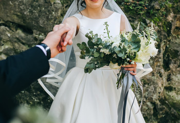 Groom is holding beautiful bride's hand in satin atlas dress outdoors in the autumn forest. Elegant wedding bouquet from eustoma , white peony and eucalyptus with grey ribbon. Happy wedding couple.