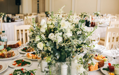 Beautifully set round tables at the restaurant. Delicious food at the wedding reception. White tablecloths and flowers decorations. Elegant space in the big hall. Happy celebration.