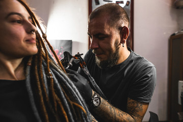 girl with dreadlocks in a tattoo parlor. The master creates a picture on the body of a young...