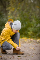 Сute child tying laces while walking in the city park on a cold autumn day. Kid boy outdors