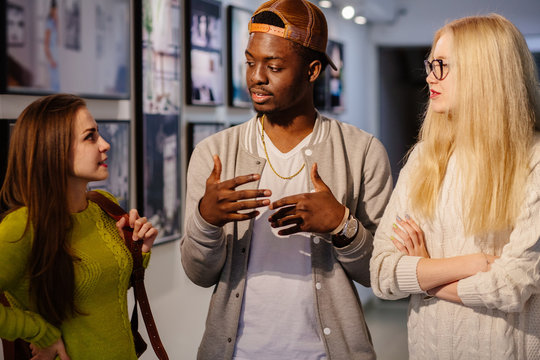 Young black hipster man talking and gesticulating with two girl friends in art photo gallery. Multi ethnic, shared values and culture friendship concept.