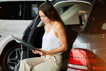 Portrait of young beautiful woman sitting on the trunk of new car while reading car properties at dealership. © alfa27
