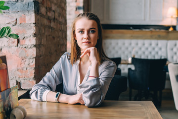 Beautiful young woman sits near big window at cafe, looking at the camera.